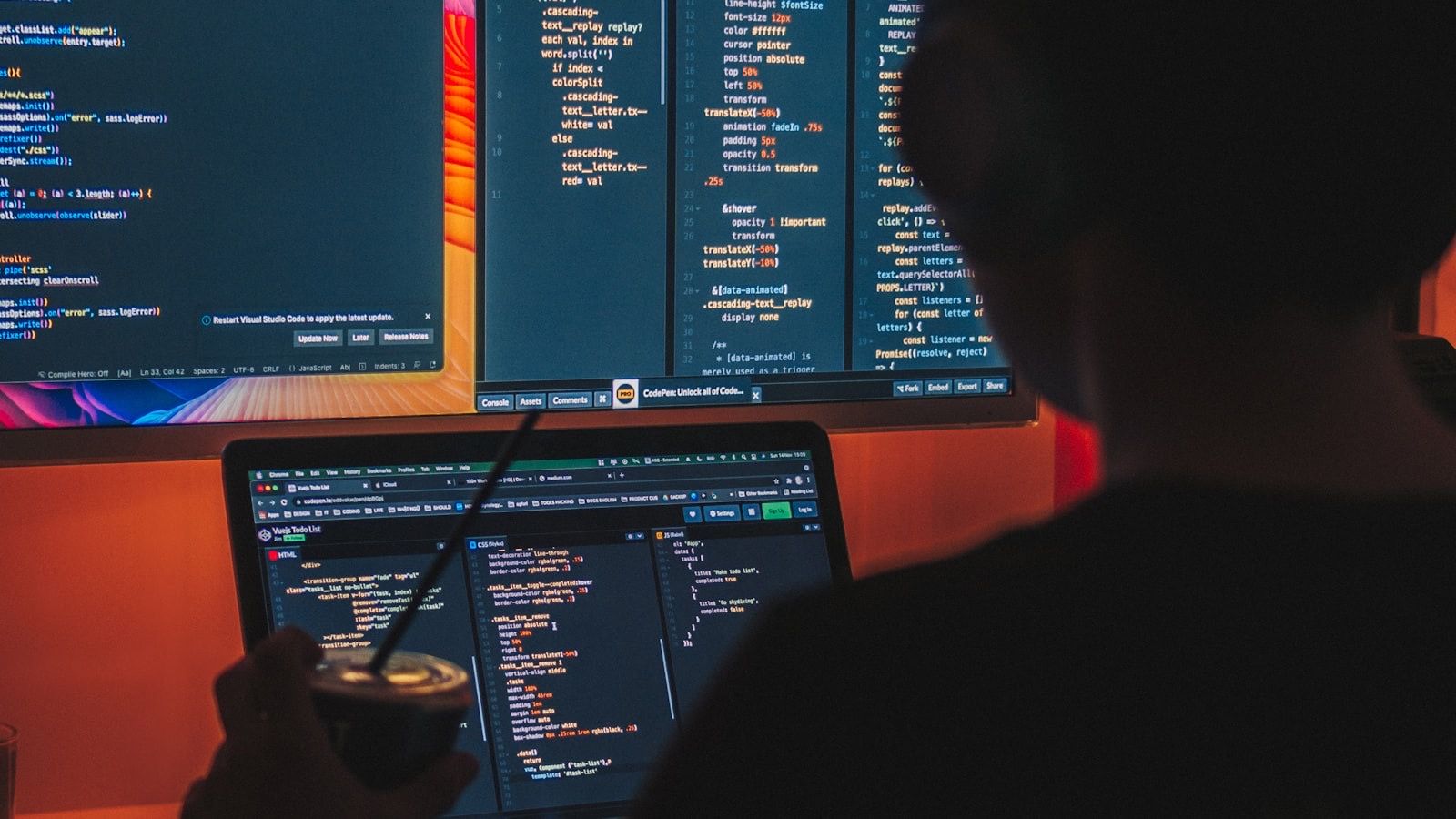 A developer at a dark desk, an editor full of code open across a monitor and a laptop.